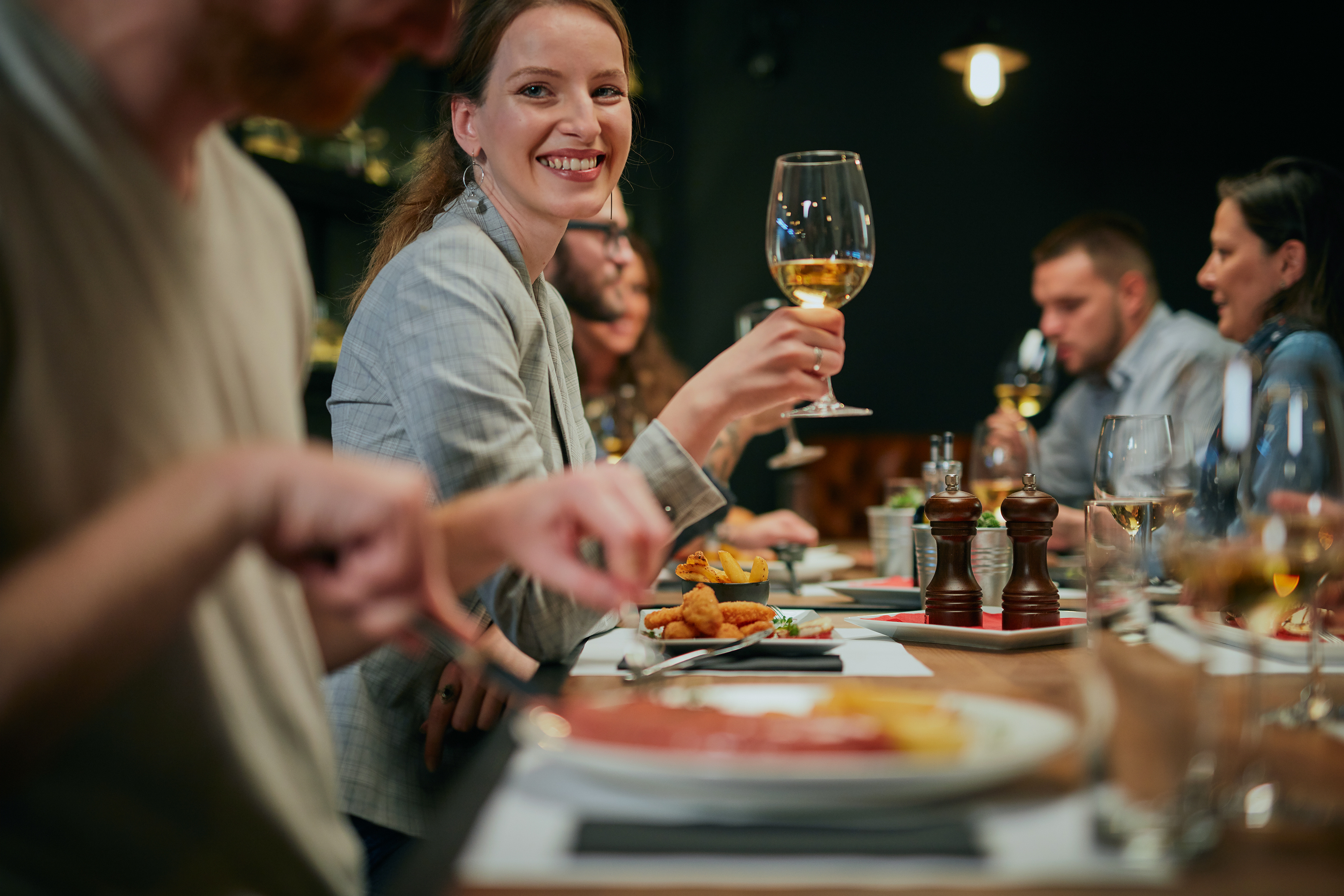 A smiling woman at a full-service restaurant holds up a glass of wine to the other guests at her table.
