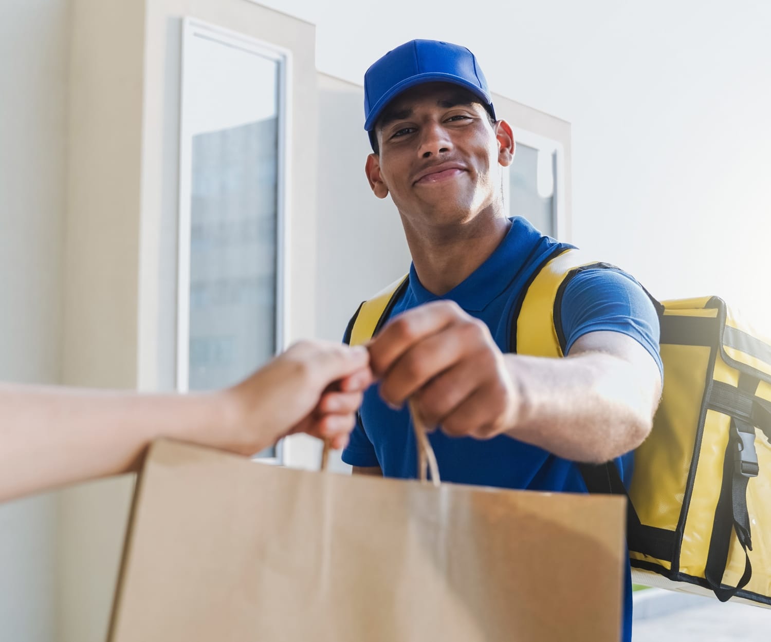 A delivery driver wearing an insulated delivery bag on one shoulder hands a customer her food.
