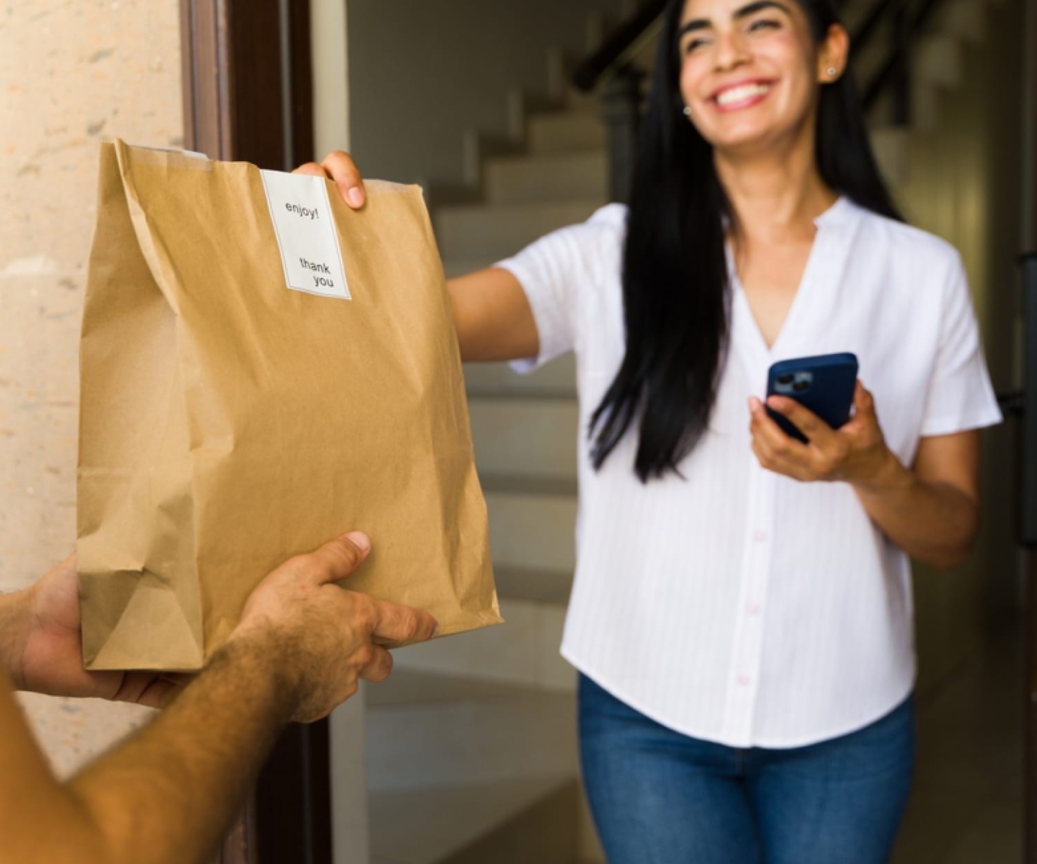 A woman stepping out of her front door holding her phone to accept a food delivery in a brown paper bag.